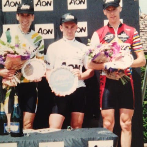 Three cyclists on a podium with AON branding, holding flowers and trophies.